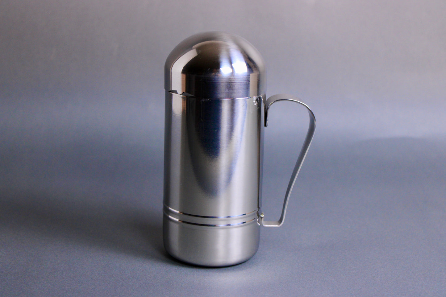cylinder shaped stainless steel oil dispenser with curved handle in grey background 