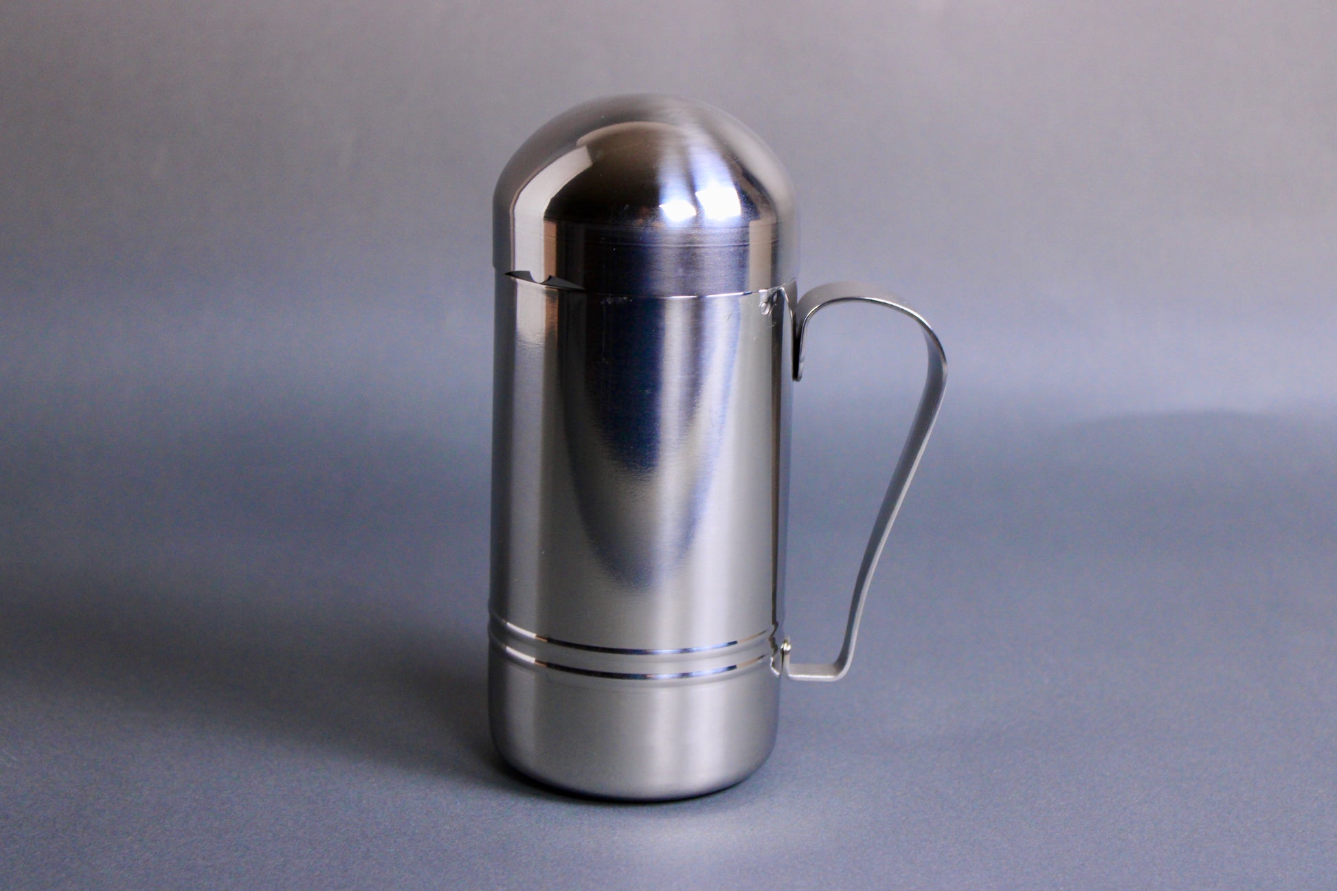 cylinder shaped stainless steel oil dispenser with curved handle in grey background 