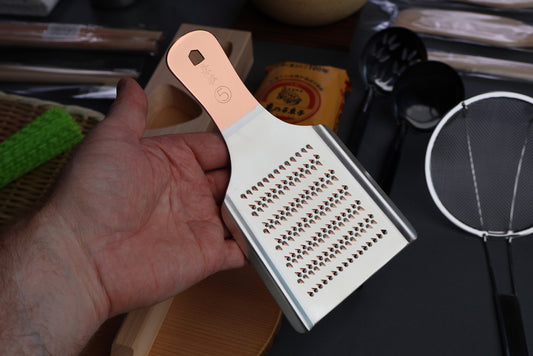 dark angled shot of hand holding copper and tin grater in front of japanese kitchenware neatly arranged black background