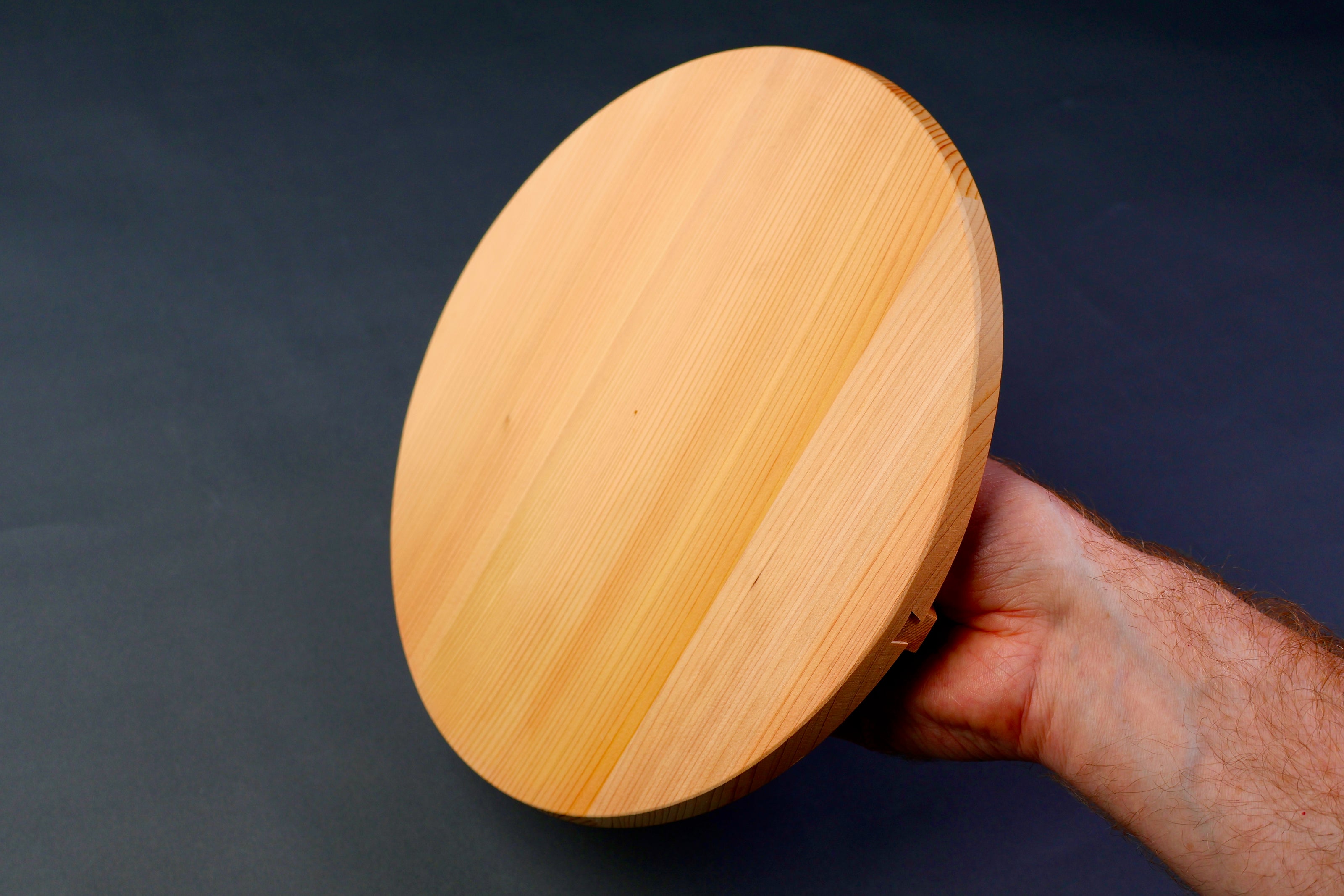 hand holding circular wooden lid yellowish hue in front of dark grey blue backdrop