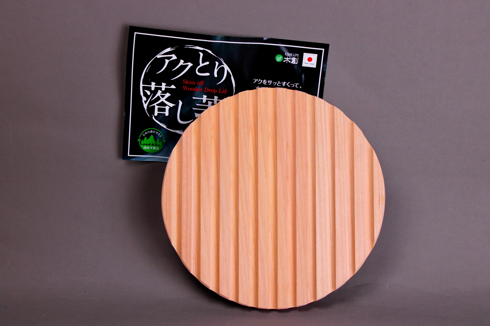 back side with ridges circular wood drop lid with long shallow handle yellowish gold hue beside black product tag with hiragana in front of grey background