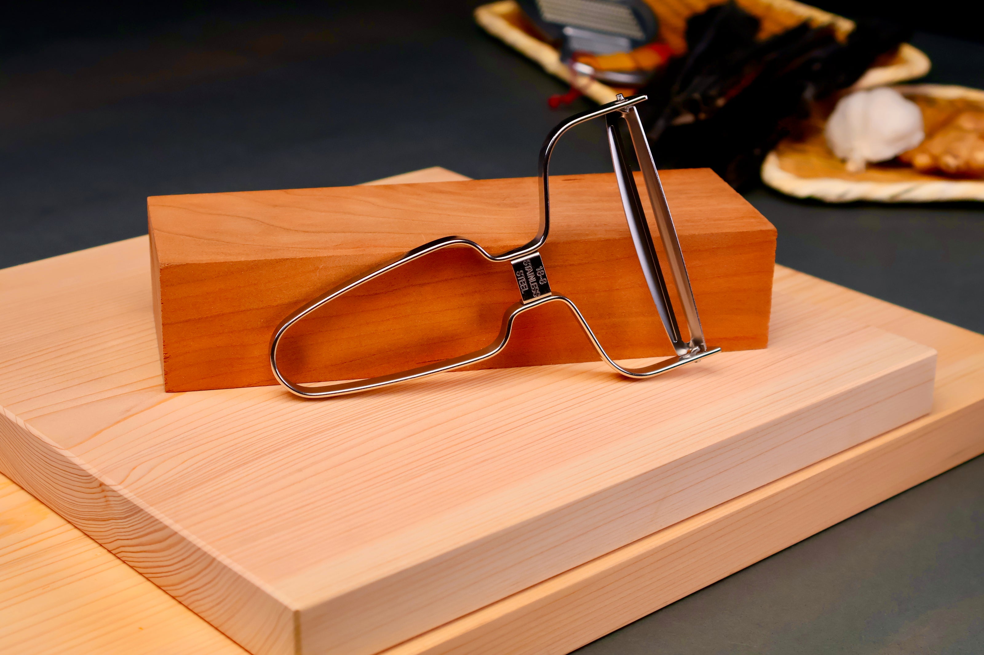 shiny angled wide vegetable peeler leaning against tan long wood block sitting atop two hinoki cutting boards in grey background