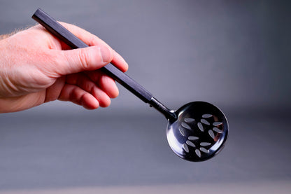hand holding a slotted steel ladle tilted to show frontal shot of holes in the metal grey background