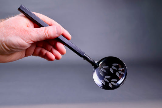 hand holding a slotted steel ladle tilted to show frontal shot of holes in the metal grey background