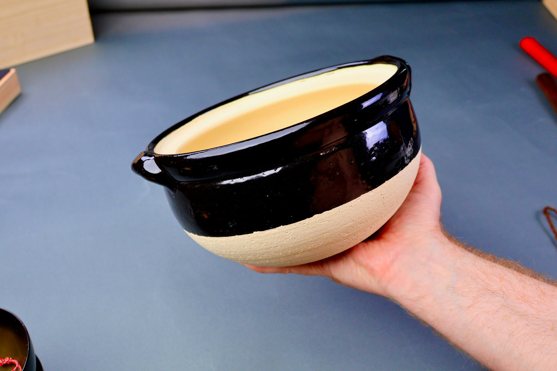 hand holding rice donabe container without lid surface is partially black glazed and unglazed grey background