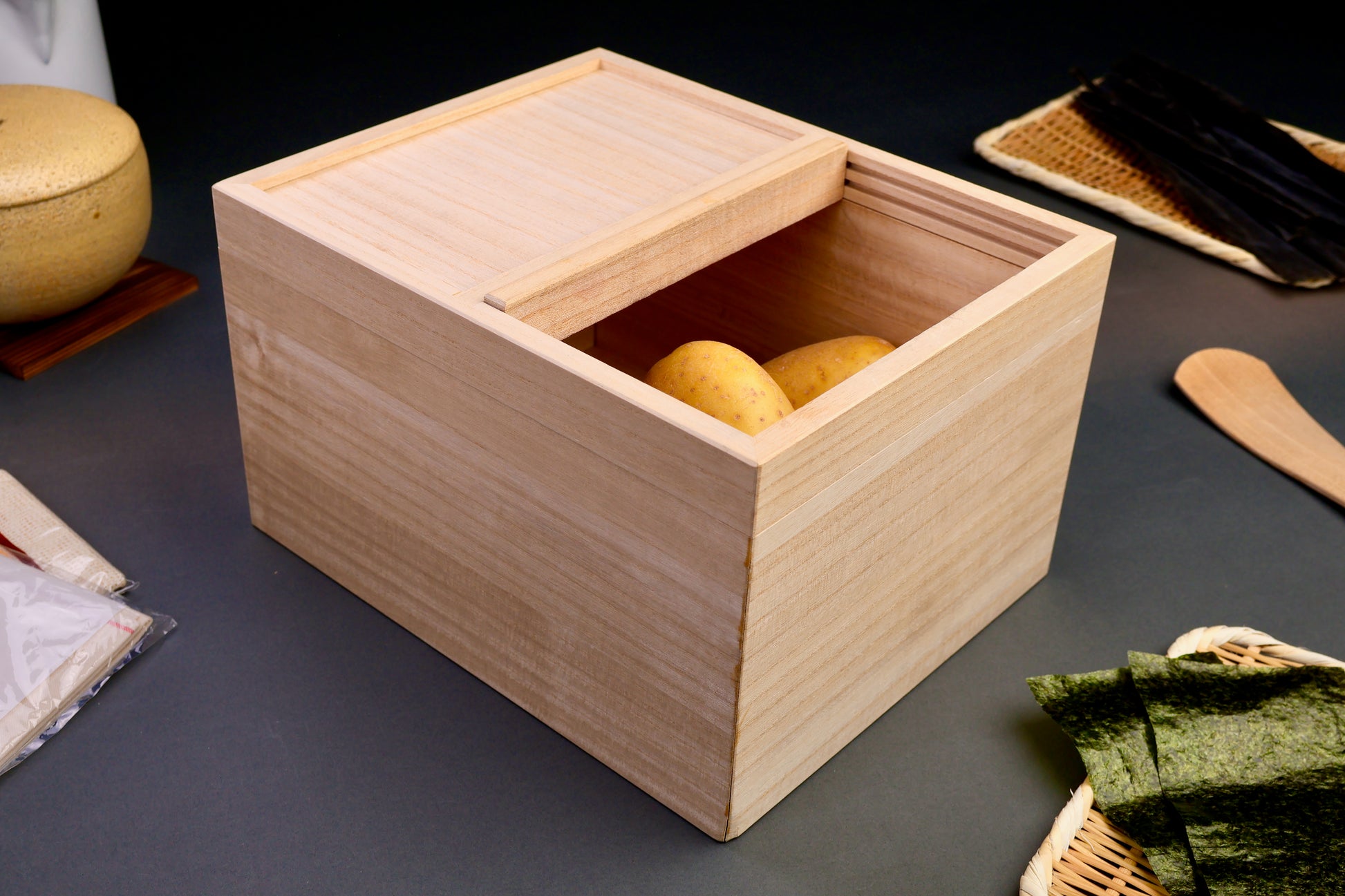 tall rectangular paulownia wood rice box sliding door on top removable with potatoes inside surrounded by japanese kitchenware dark grey background