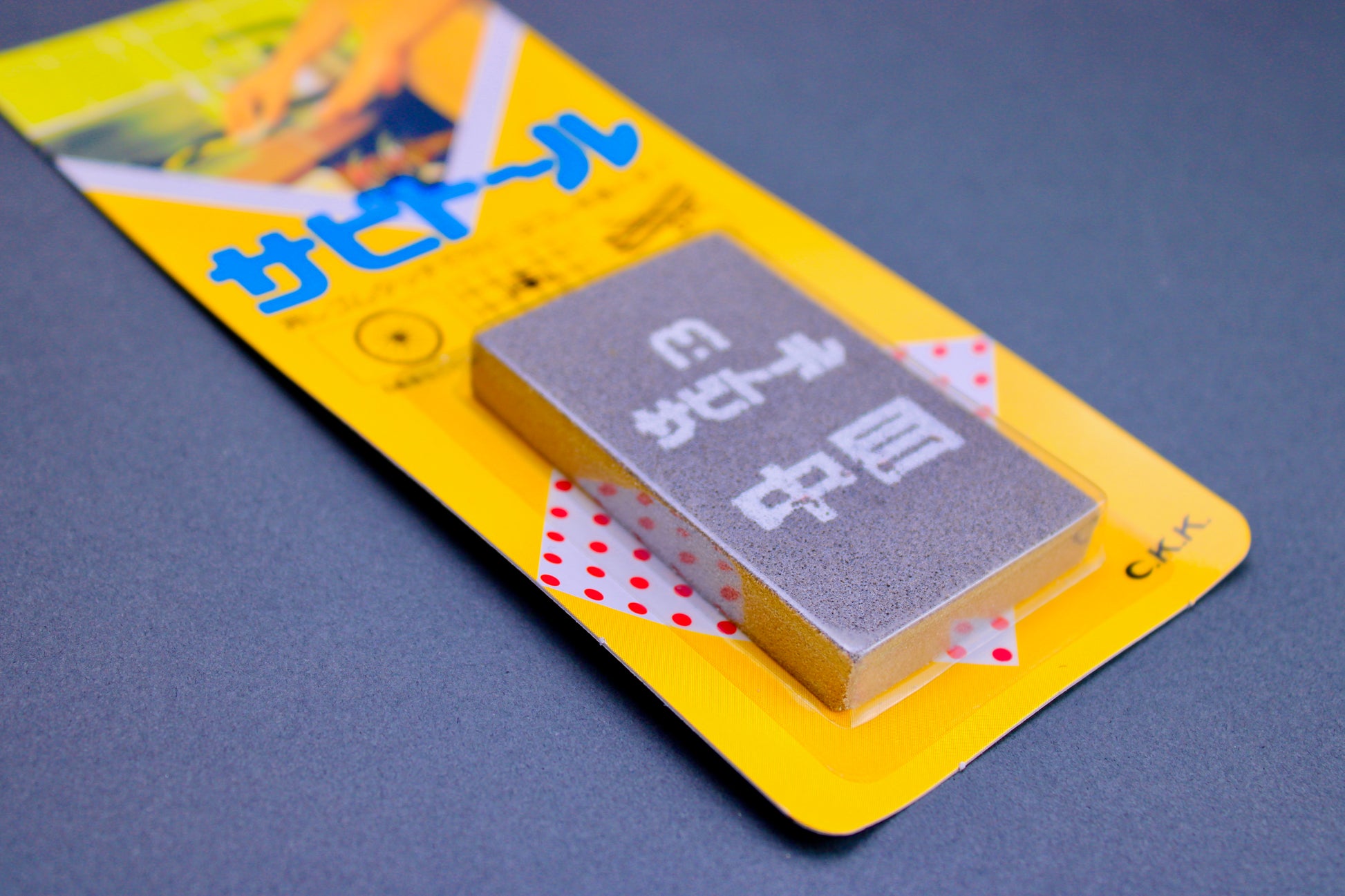 close up of grey rectangular rust eraser inside clear plastic with bright yellow product card with red dots and blue hiragana on grey background