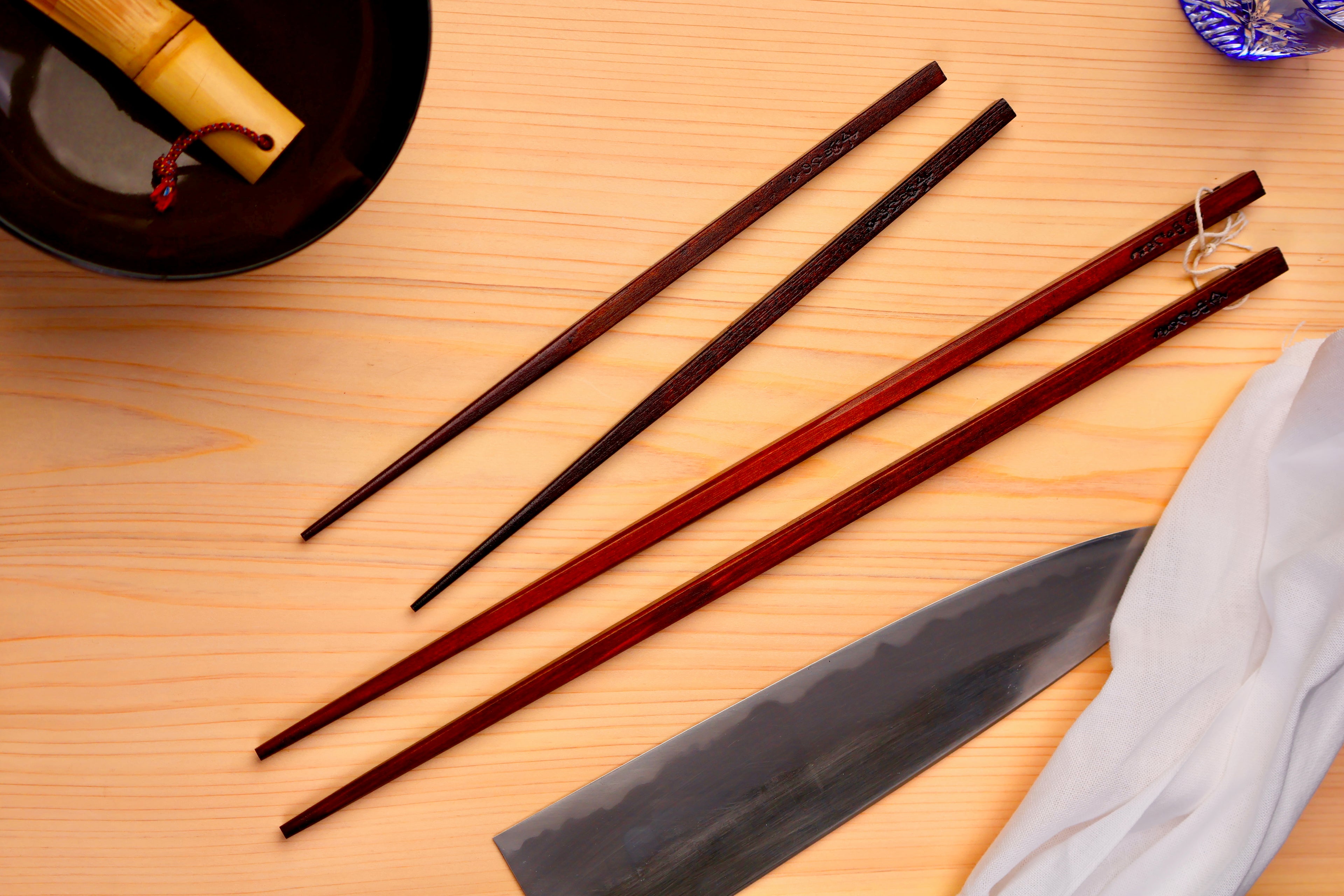 two dark brown painted hinoki wood chopsticks on top of hinoki wood cutting board surrounded by kitchen knife white towel black bowl with and bluish clear glass 