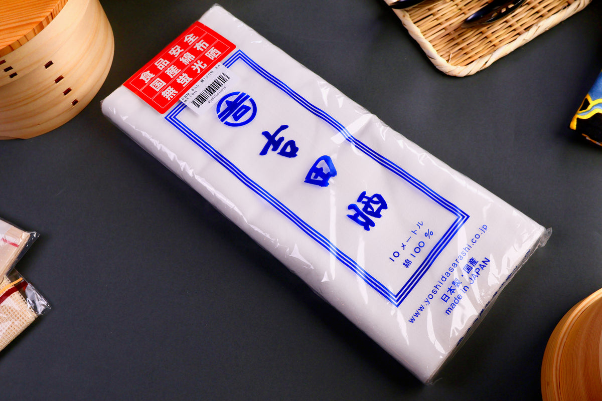 bright white long rectangular sarashi towel in clear plastic packaging with red product card on top with kanji and blue product printing surrounded by japanese kitchenware on gray backdrop 