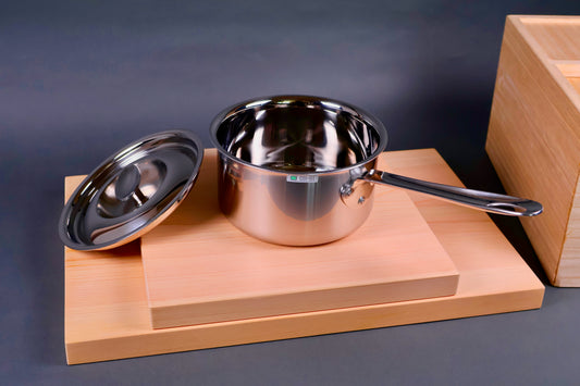 closeup shot of japanese steel 16cm saucepan with curved rim and curved lid beside items sit atop wood boards beside wood rice box grey background