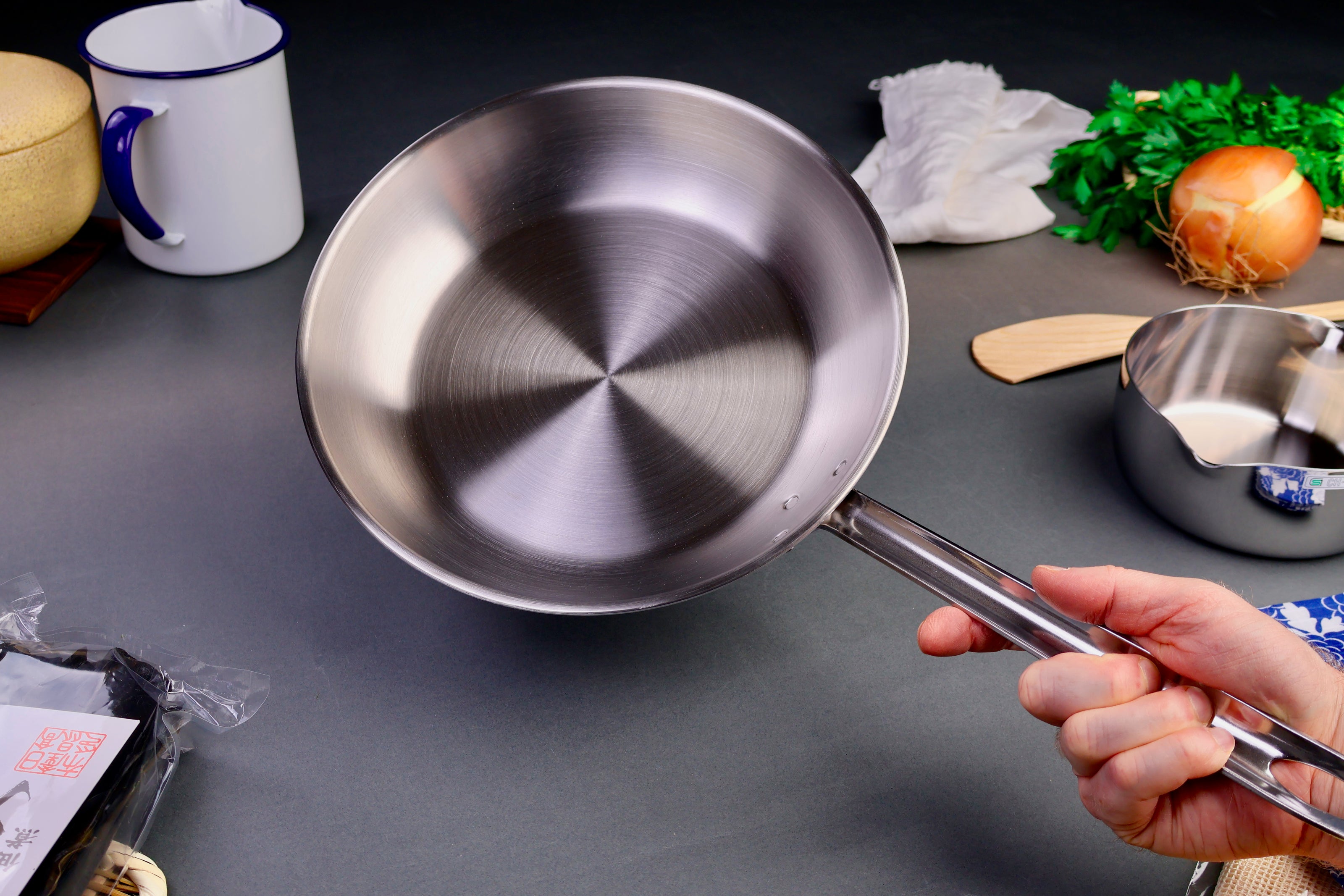 hand holding saute pan with long handle shiny steel surrounded by japanese kitchenware grey background