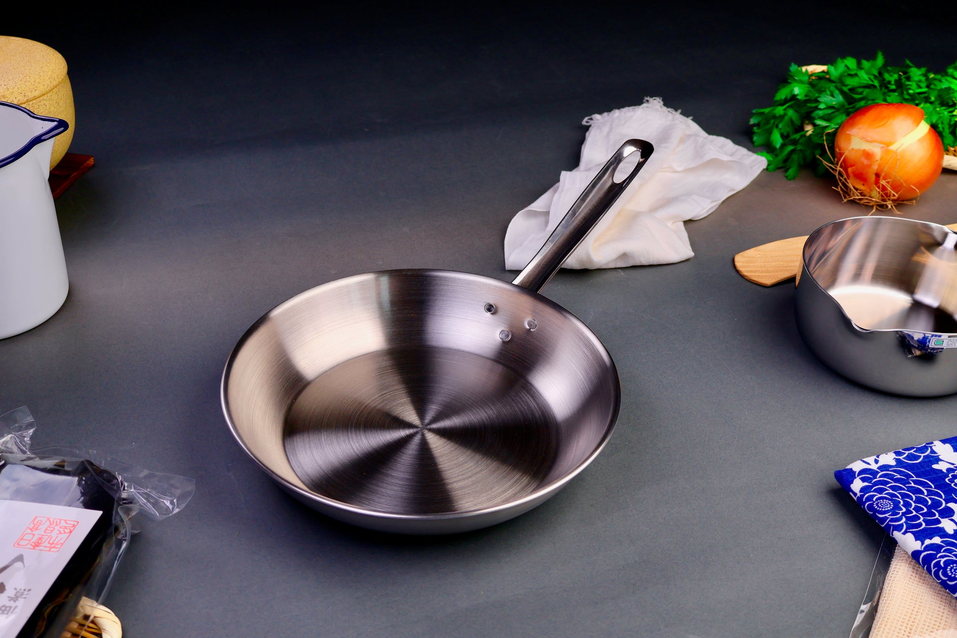 high quality stainless steel saute pan with long elegant handle surrounded by Japanese kitchenware with light grey background
