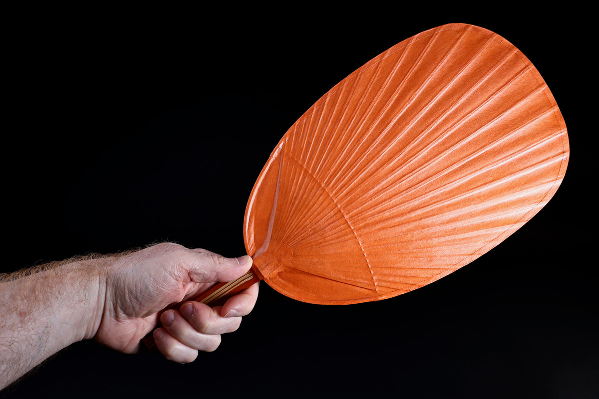 Shibu grilling fan orange held in hand against black background, showing full oval fan face and bamboo handle