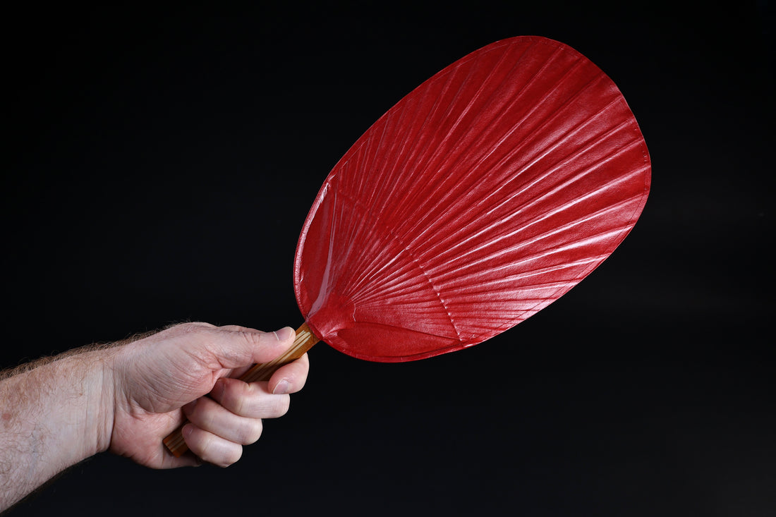Shibu grilling fan red held in hand against black background, showing full oval fan face and bamboo handle