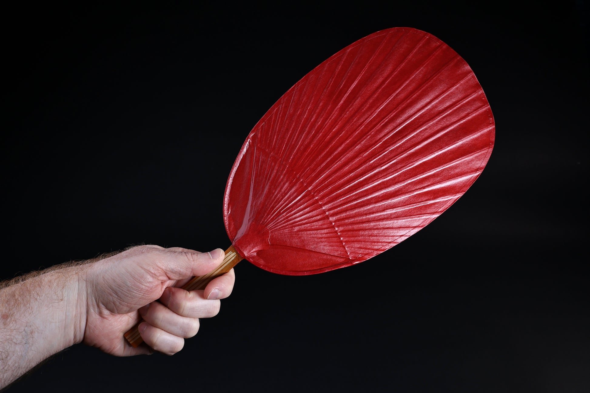 Shibu grilling fan red held in hand against black background, showing full oval fan face and bamboo handle