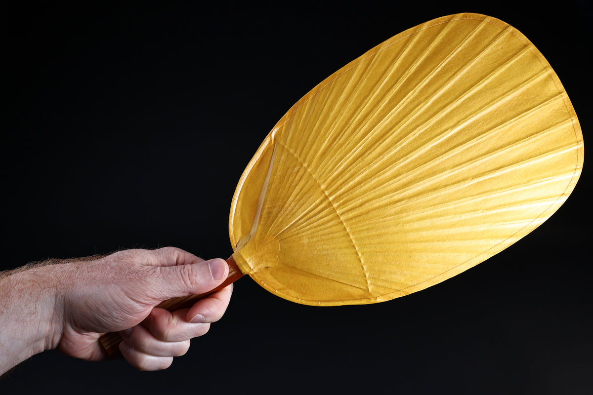 Shibu grilling fan yellow held in hand against black background, showing full oval fan face and bamboo handle