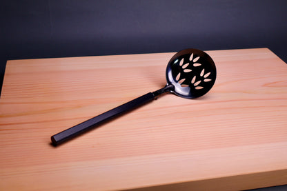 slotted steel ladle darkened with oxidized finish laying atop hinoki wood cutting board in grey backdrop