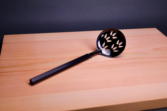 slotted steel ladle darkened with oxidized finish laying atop hinoki wood cutting board in grey backdrop