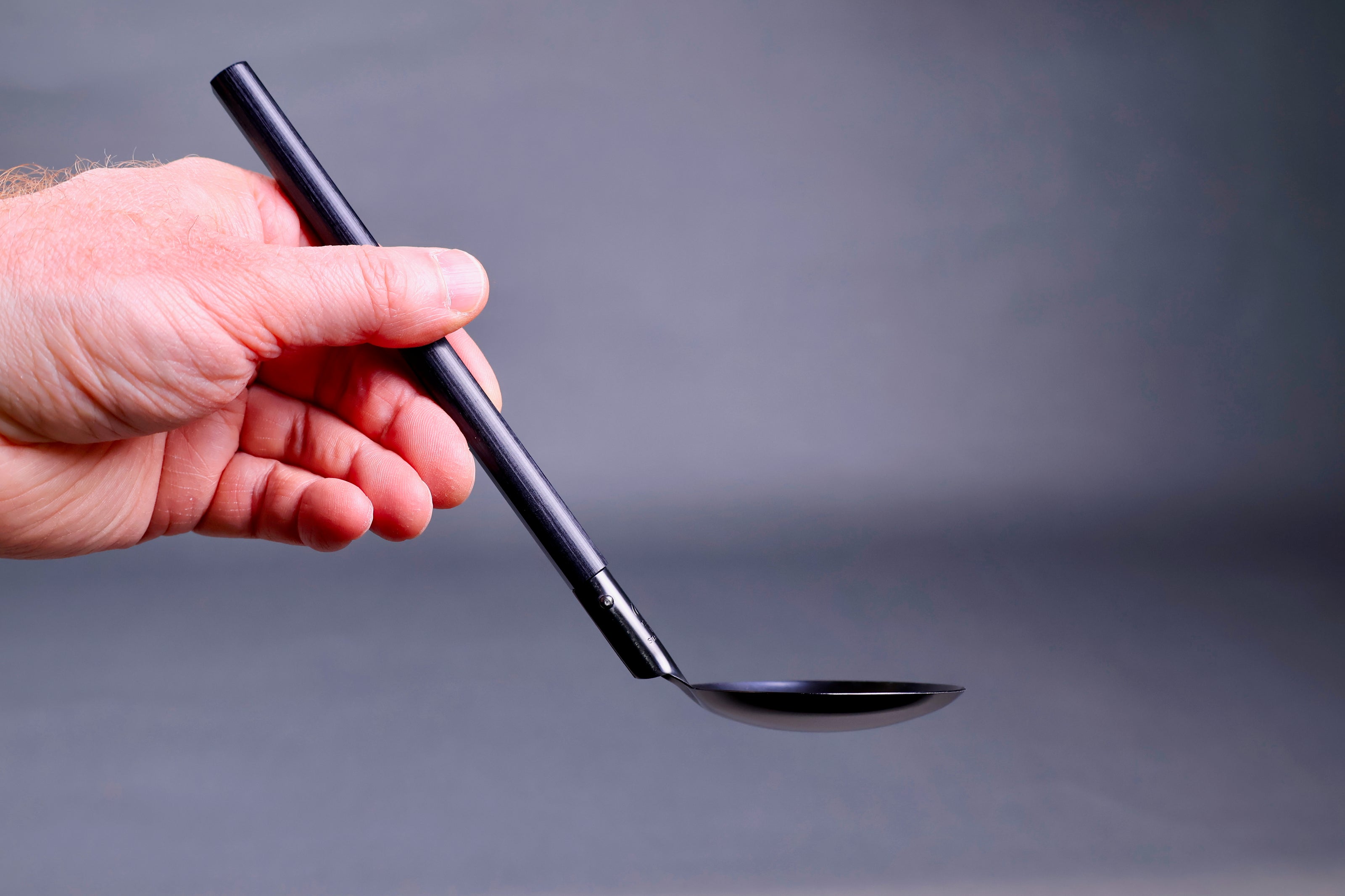 hand holding darkened steel ladle with angled wood handle in grey background
