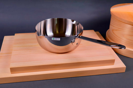 shiny silver metal pan with long handle on wood boards beside round wood steamer in grey background