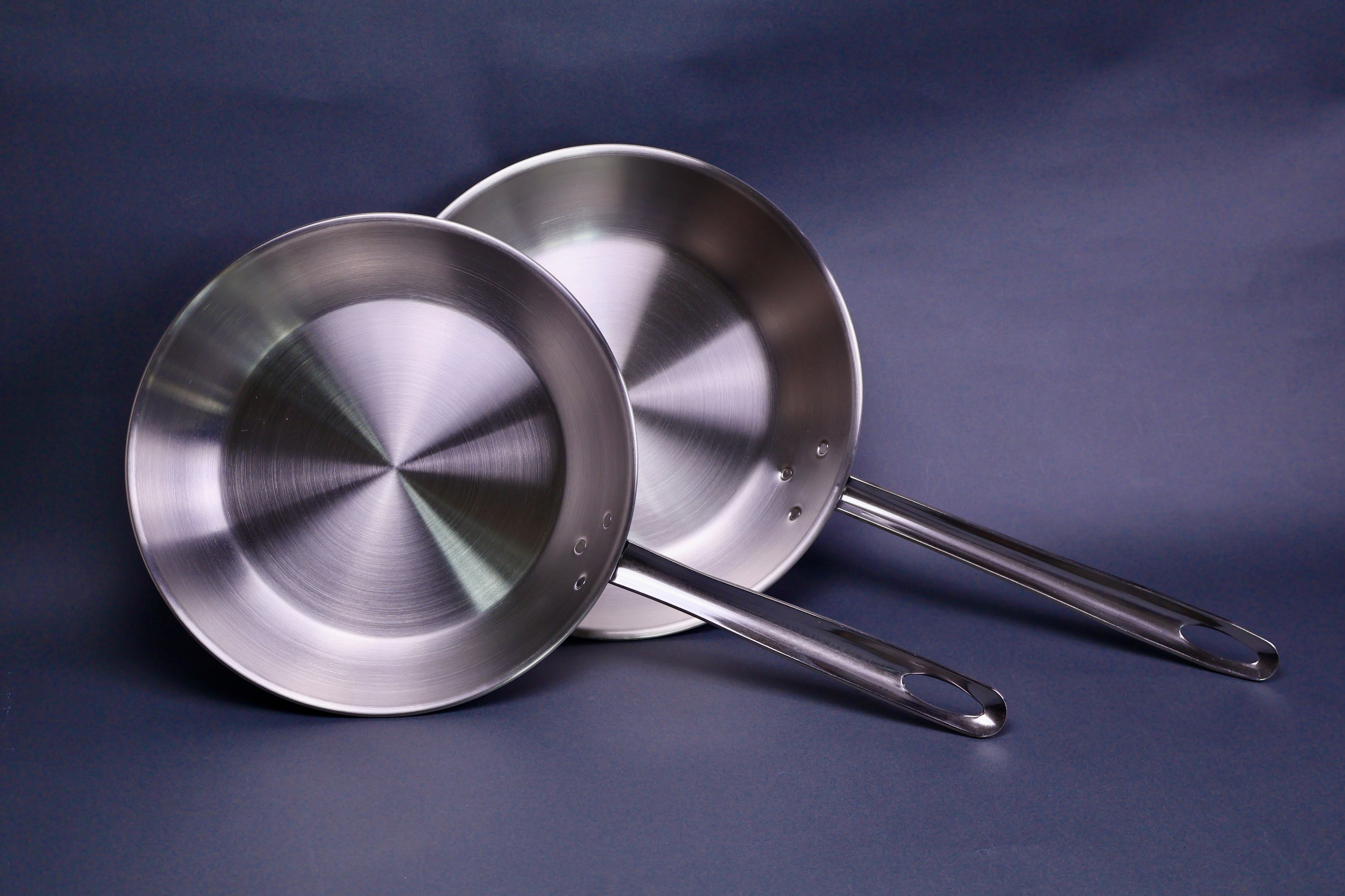 two stainless steel round dark shiny silver pans each with long straight handles with loop holes in bluish grey backdrop