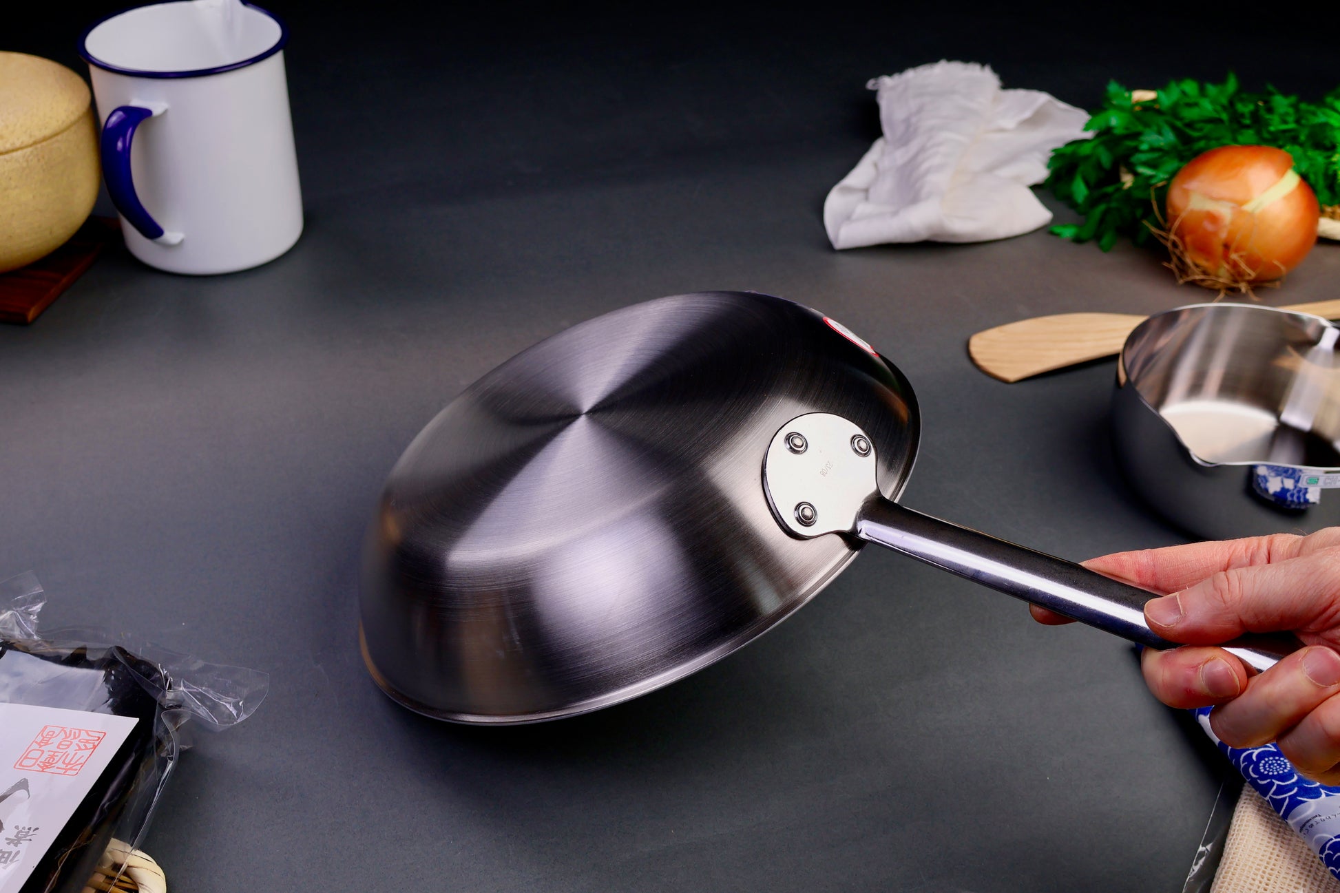 underside of shiny elegant saute pan with smooth rivets holding the handle onto the pan with dark grey backdrop with kitchenware