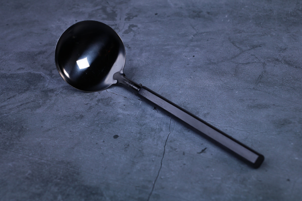 Stainless steel oxidized ladle — blackened solid bowl and octagonal wooden handle on concrete surface