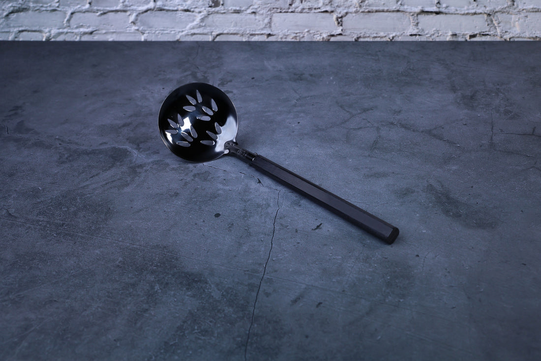 Stainless steel oxidized perforated ladle — full view showing blackened bowl, perforations, and wooden handle