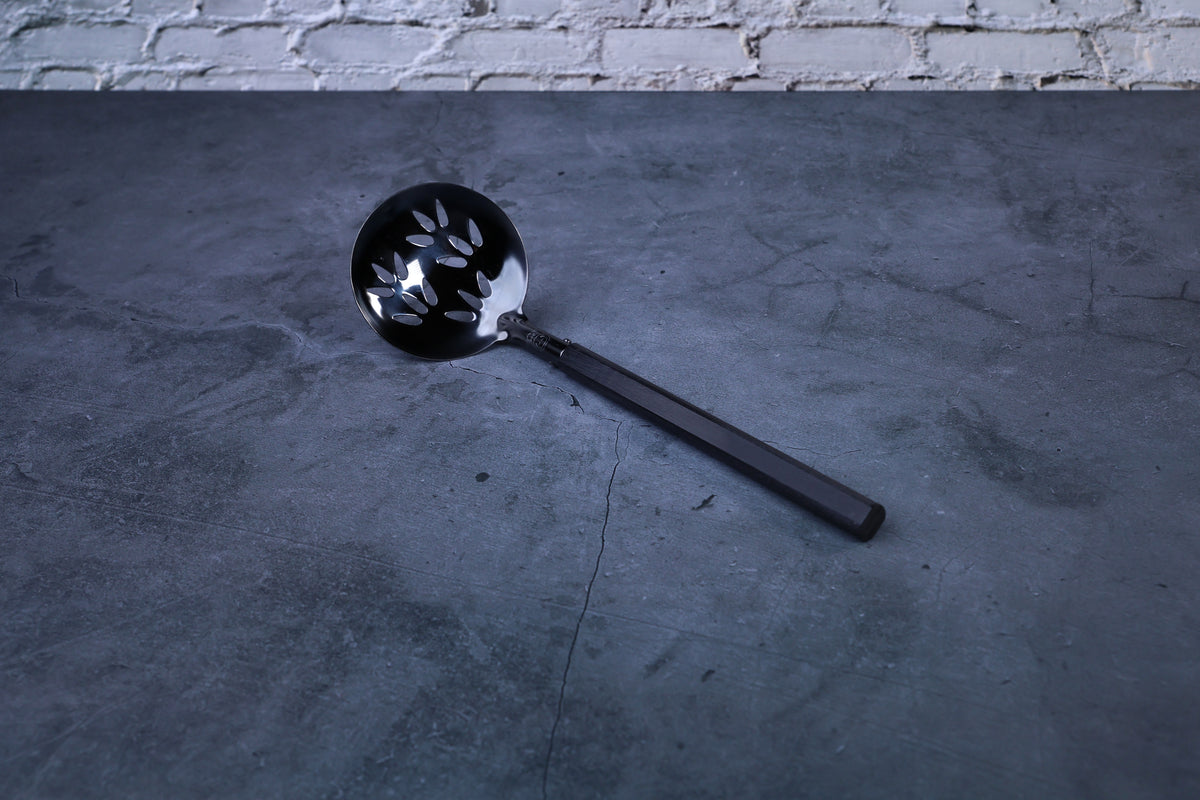 Stainless steel oxidized perforated ladle — full view showing blackened bowl, perforations, and wooden handle
