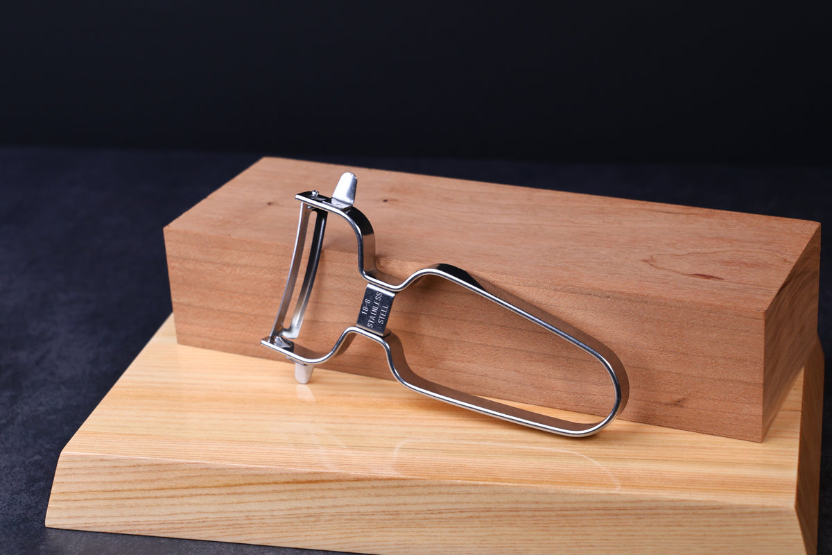 Stainless steel vegetable peeler — full view showing swivel blade, wire handle, and eye remover on wood surface