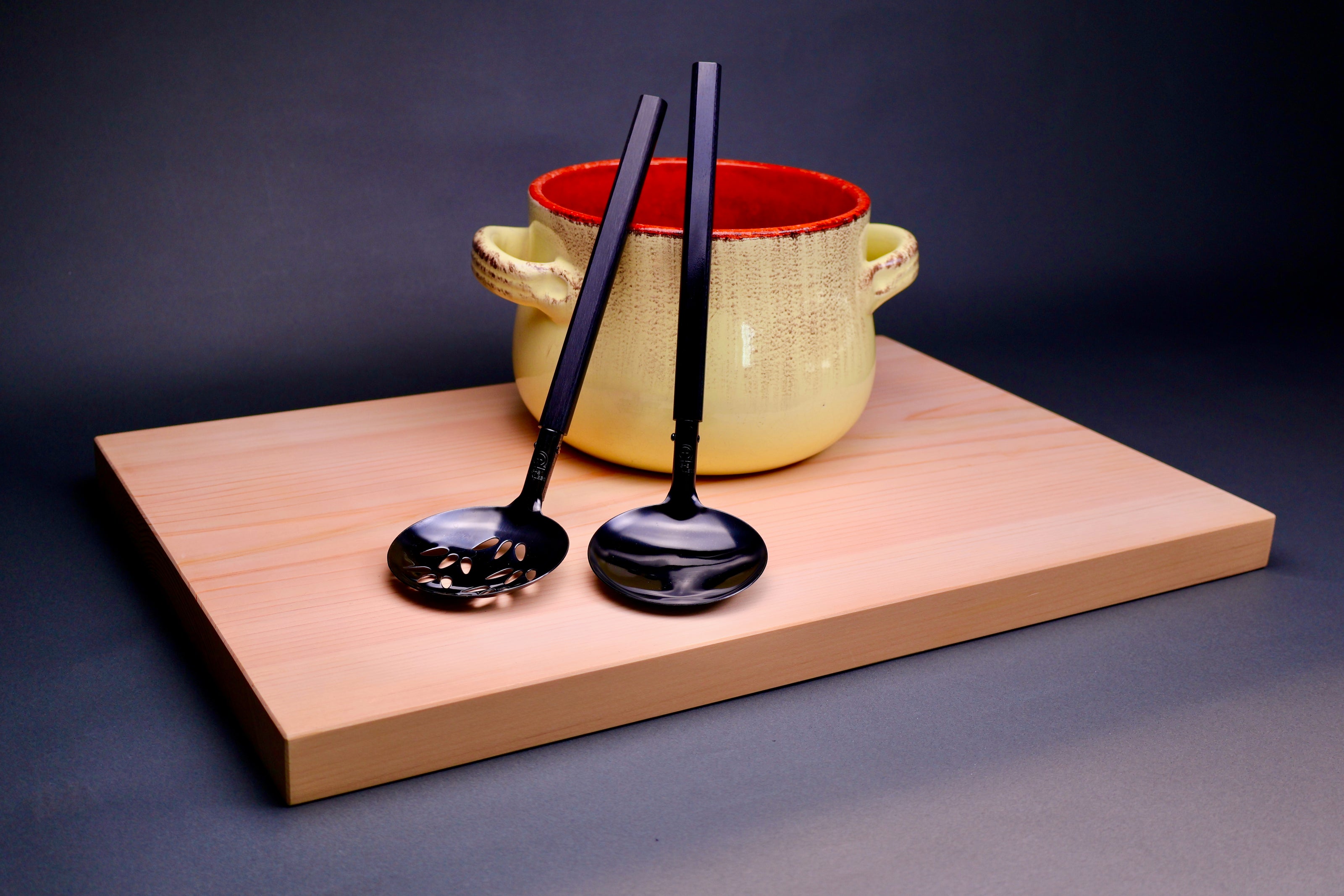 two ladles slotted and solid leaning against ceramic yellowish reddish pot atop hinoki cutting board in grey backdrop