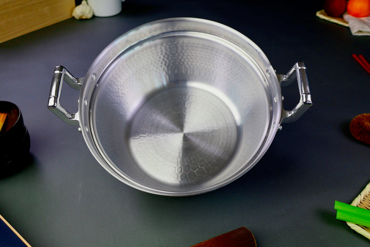 interior of two handled stepped aluminum silvery pot with shiny hammered surface surrounded by japanese kitchenware on a dark grey backdrop 