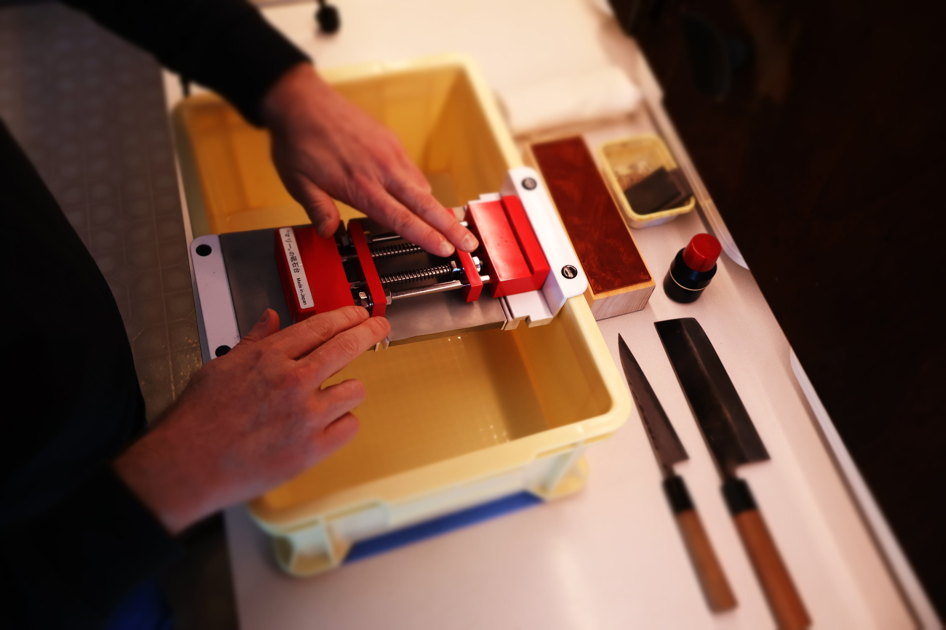 kitchen counter with tall yellow container holding suehiro sliding rack with white edges atop sits marie whetstone holder red and two hands moving the product in place surrounded by knives 
