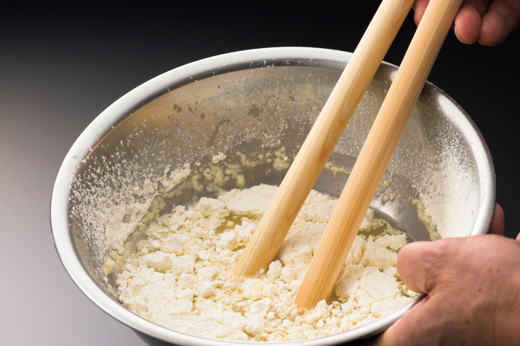 hand holding shiny bowl containing white partially mixed tempura batter other hand holding thick saibashi round chopsticks mixing batter in dark background