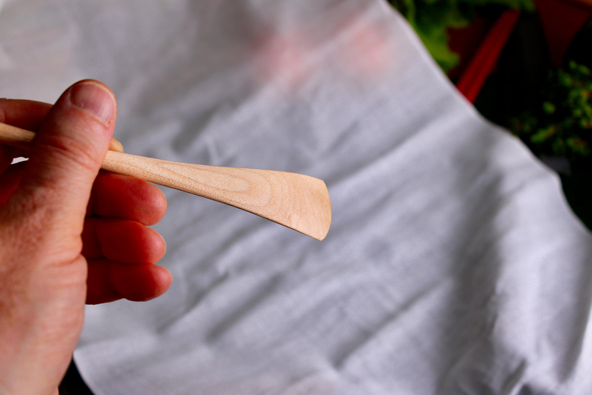 hand holding a long thin wooden tan jam spoon showing wood grain on white cloth background with kitchenware