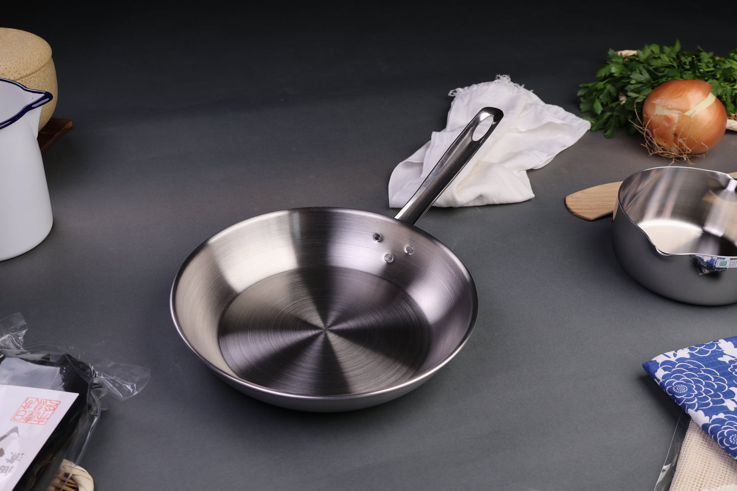 three layer aluminum clad stainless steel saute pan with long curved handled silver tone centered around japanese kitchenware dark grey backdrop