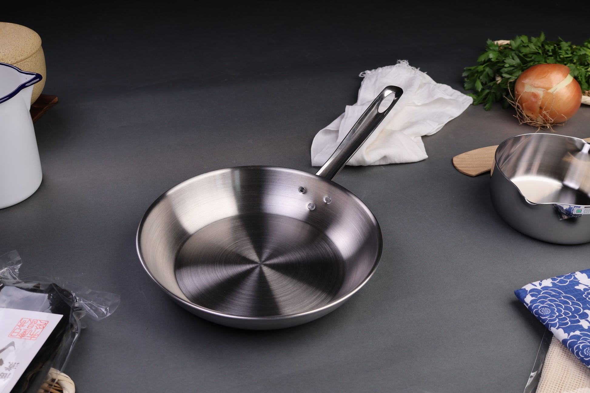three layer aluminum clad stainless steel saute pan with long curved handled silver tone centered around japanese kitchenware dark grey backdrop