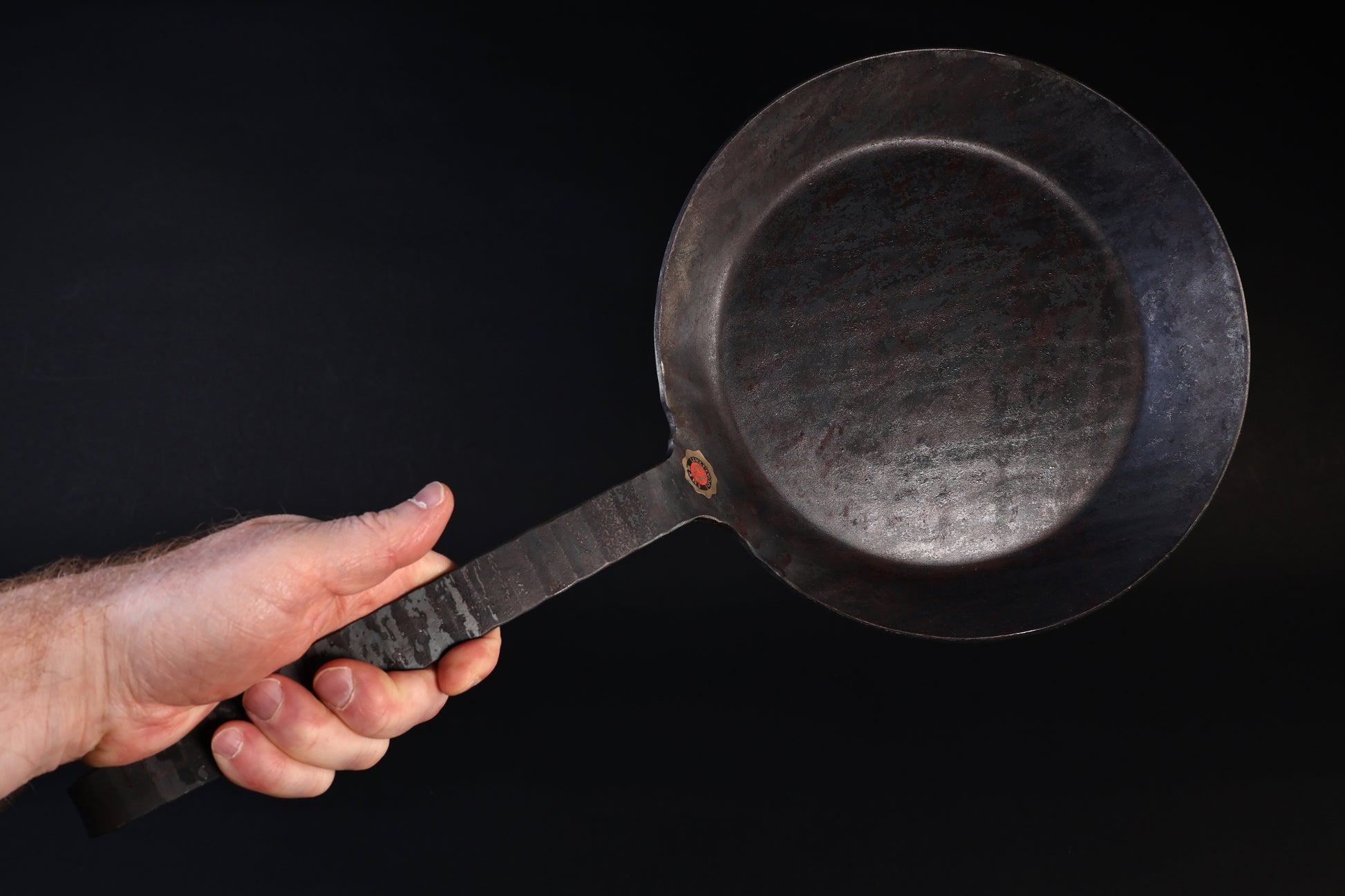 Turk hand-forged iron frying pan held by the handle against a dark background, cooking surface facing forward