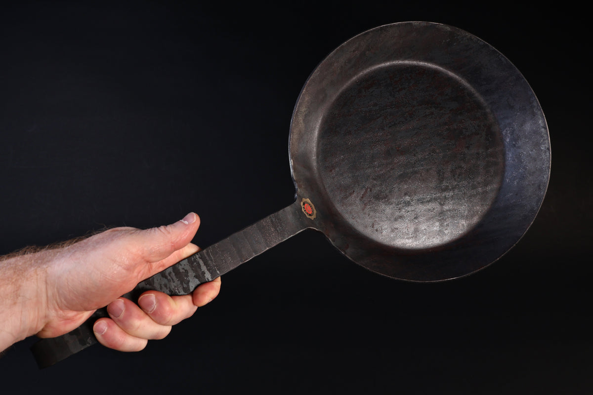 Turk hand-forged iron frying pan held by the handle against a dark background, cooking surface facing forward