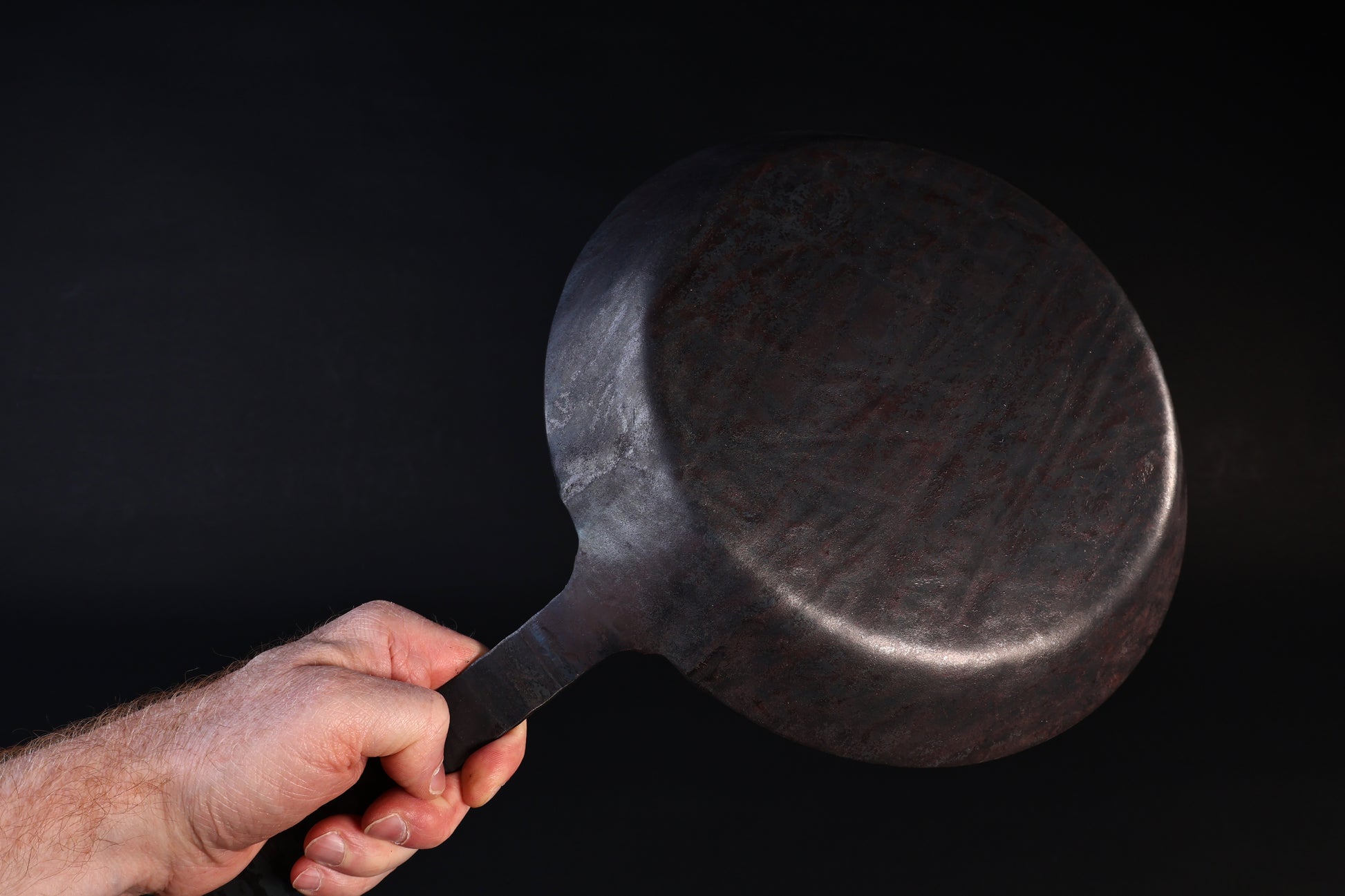 Turk hand-forged iron frying pan held by the handle showing the hammered underside against a dark background
