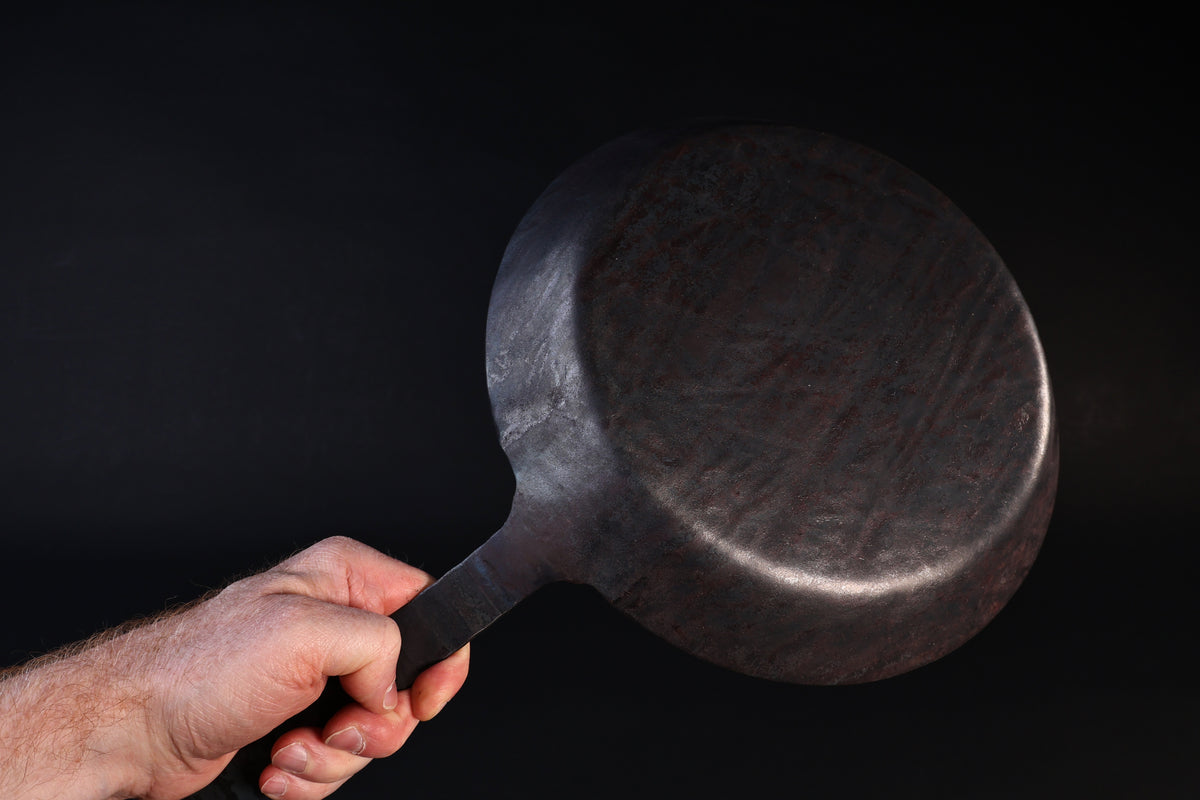 Turk hand-forged iron frying pan held by the handle showing the hammered underside against a dark background