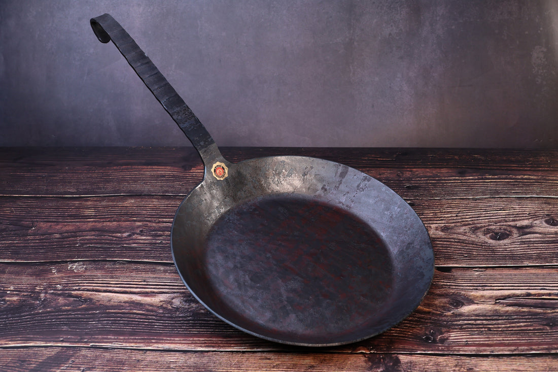 Turk hand-forged iron frying pan with its long hanging-hook handle and brass quality seal, seen from the front