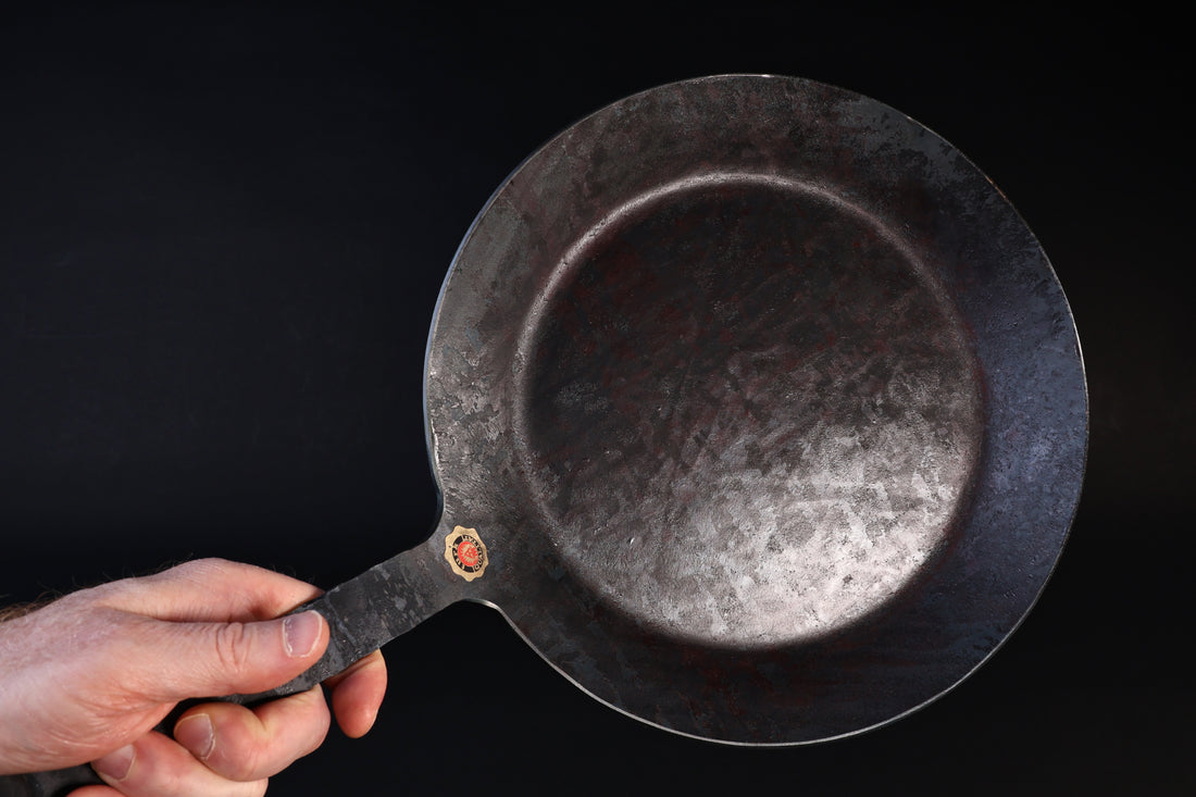 Turk hand-forged iron frying pan held by the handle against a dark background, cooking surface facing forward
