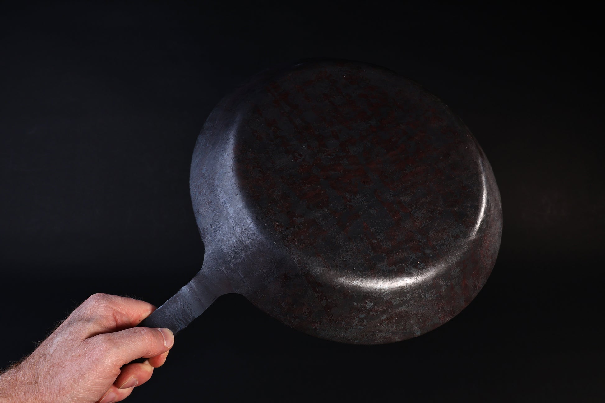 Turk hand-forged iron frying pan held by the handle showing the hammered underside against a dark background