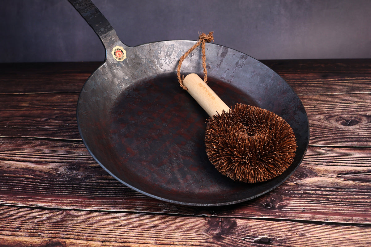 Turk hand-forged iron frying pan shown with a palm-fiber tawashi cleaning brush resting in the bowl