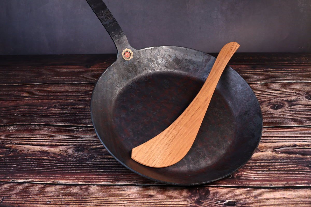 Turk hand-forged iron frying pan with a wooden cooking turner resting inside the bowl