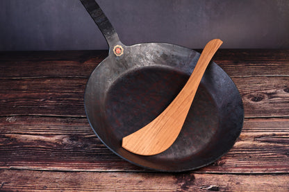 Turk hand-forged iron frying pan with a wooden cooking turner resting inside the bowl