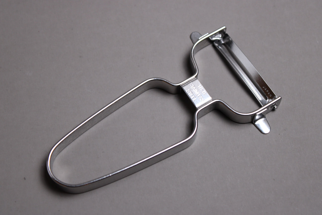 stainless steel vegetable peeler with curved blade wide grip shown on grey background angled shot of peeler
