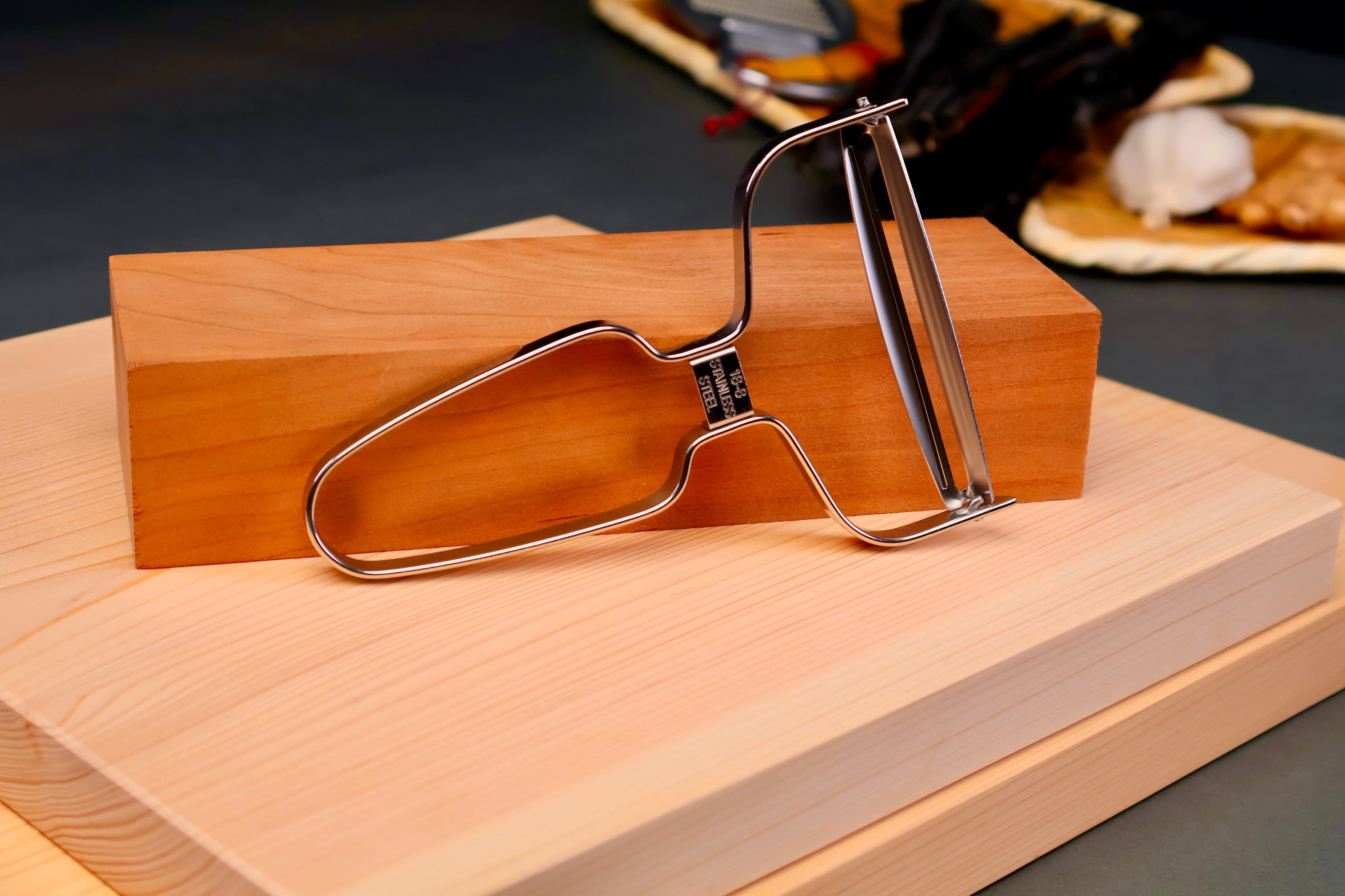 shiny angled wide vegetable peeler leaning against tan long wood block sitting atop two hinoki cutting boards in grey background