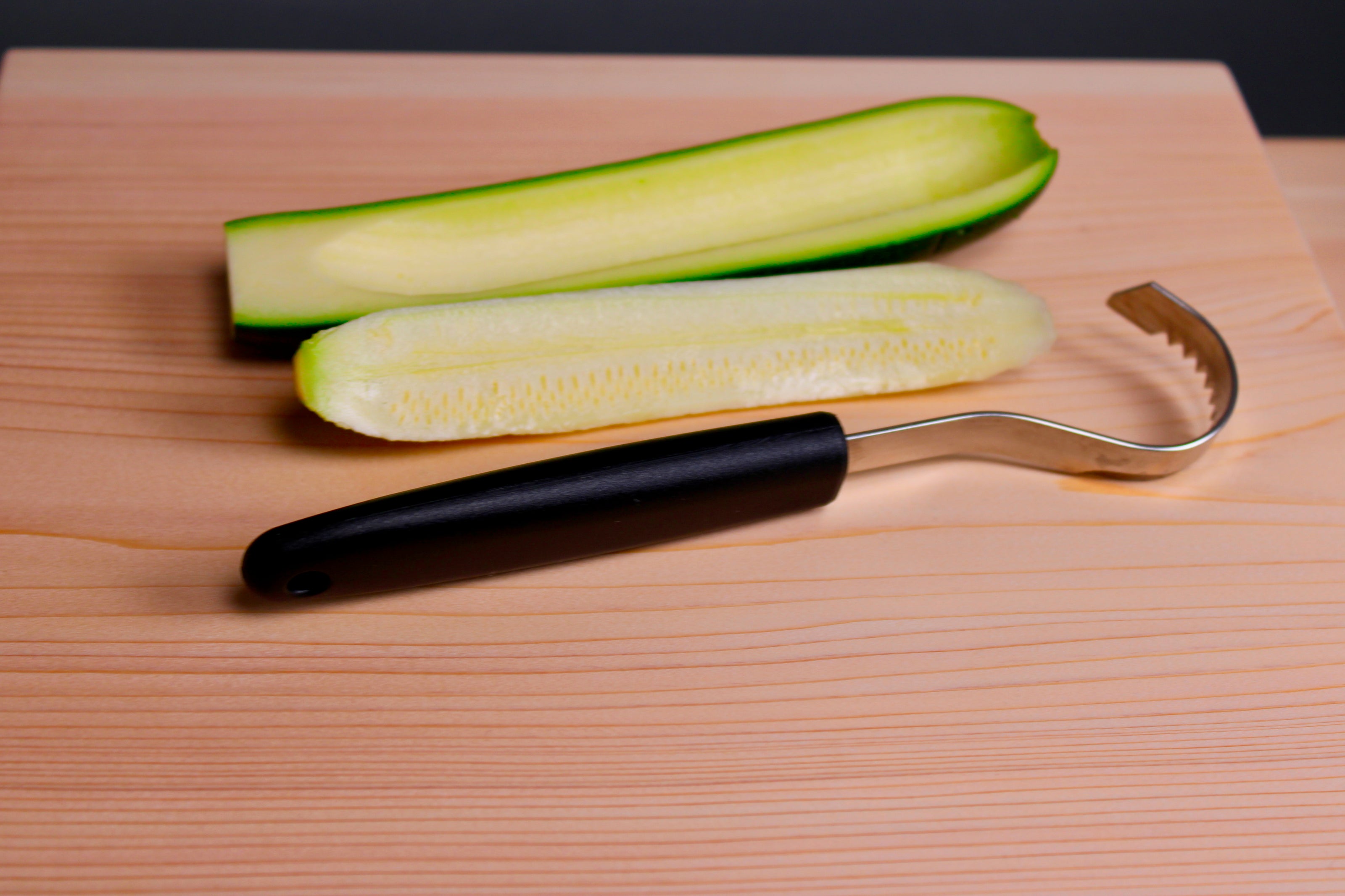 long thin black plastic handled stainless steel curved serrated tool butter curler beside cored zucchini atop wood cutting board